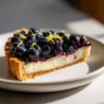 A close-up of a slice of blueberry mascarpone tart, topped with fresh blueberries and lemon zest.