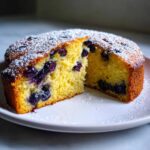 A slice of moist blueberry orange olive oil cake dusted with powdered sugar on a white plate.