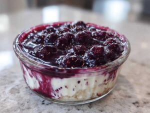 A close-up of a Blueberry Vanilla Bean Panna Cotta served in a clear glass bowl, topped with a generous blueberry compote.