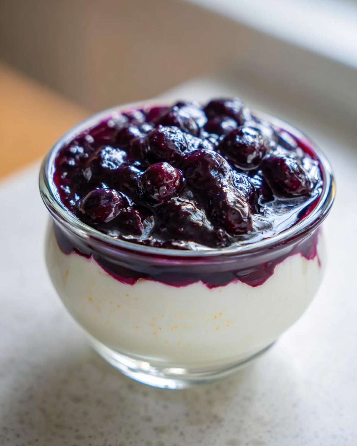 A close-up of a creamy Blueberry Vanilla Bean Panna Cotta topped with a rich blueberry compote in a glass dish.