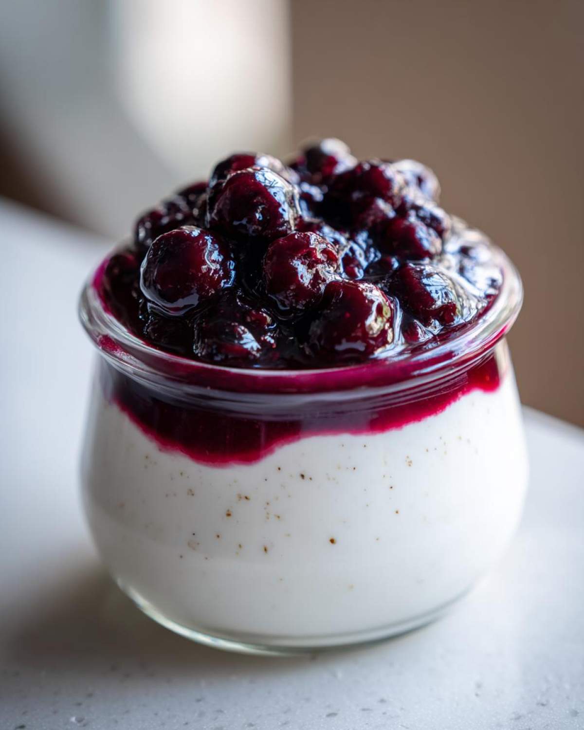 Close-up of a creamy Blueberry Vanilla Bean Panna Cotta in a glass jar, topped with fresh blueberries and sauce.