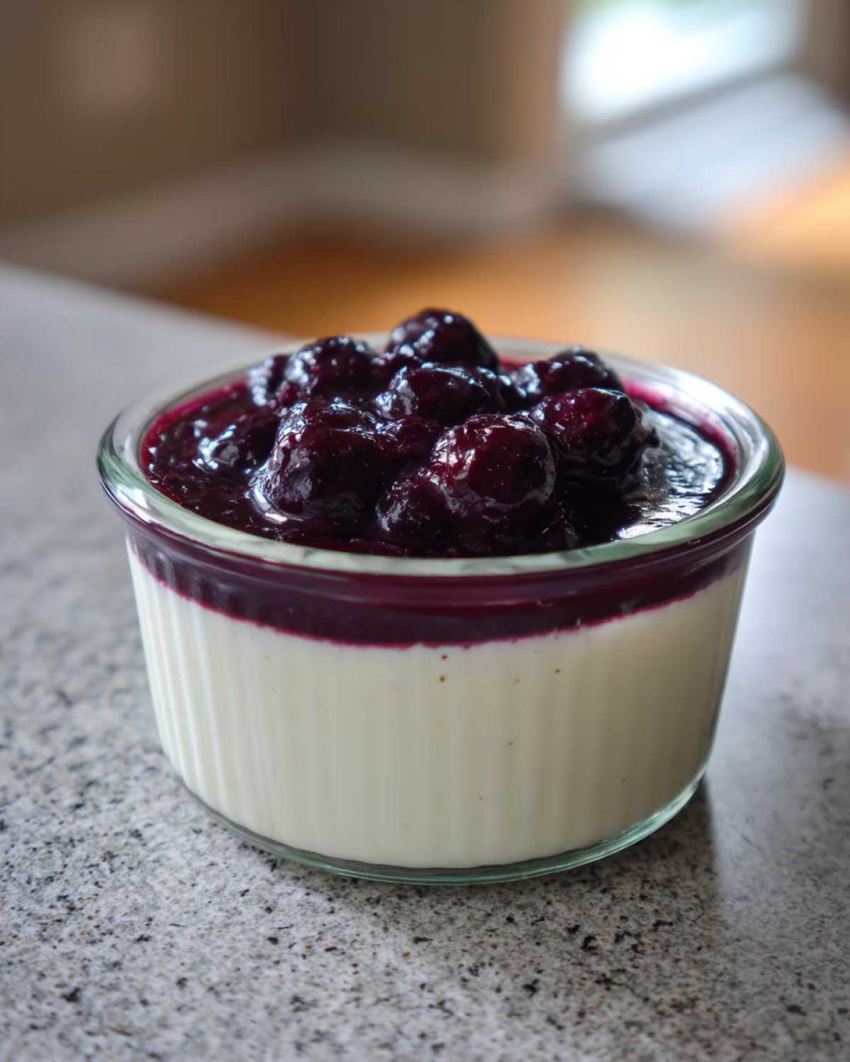A close-up of a creamy Blueberry Vanilla Bean Panna Cotta topped with fresh blueberries and sauce in a glass ramekin.