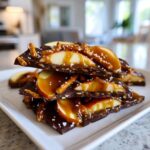 A stack of dark chocolate caramel apple bark pieces, topped with fresh apple slices, pretzels, and a drizzle of caramel.