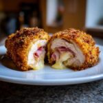 A close-up of a crispy Chicken Cordon Bleu, cut in half, revealing melted cheese and ham inside.