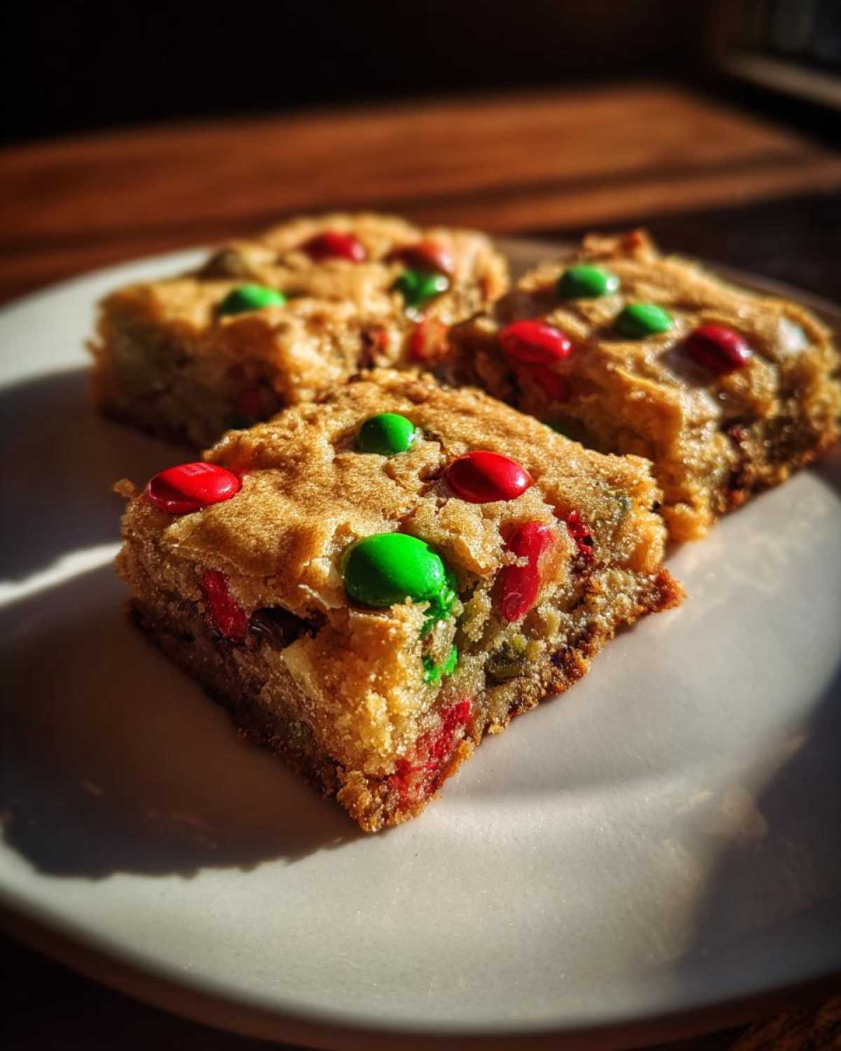 Close-up of three Christmas cookie bars topped with red and green M&Ms on a white plate.