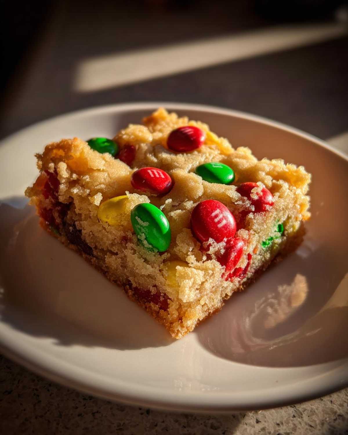 A slice of Christmas cookie bars topped with red, green, and yellow M&Ms on a white plate.