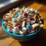 A close-up of a blue bowl filled with a colorful Christmas snack mix, featuring pretzels, cereal squares, popcorn, and candies.