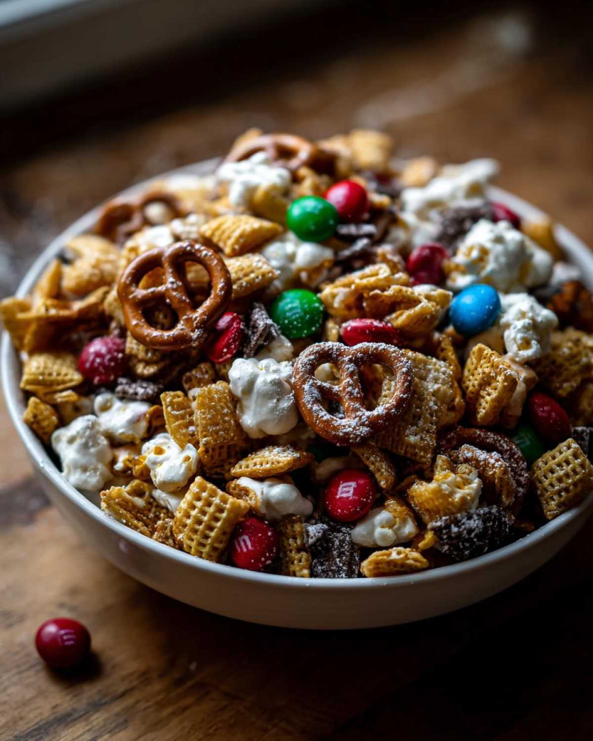 A white bowl overflowing with a delicious Christmas snack mix, featuring pretzels, cereal squares, popcorn, and colorful M&Ms.