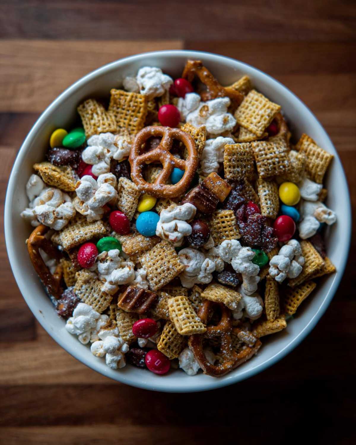A close-up overhead view of a white bowl filled with a colorful Christmas snack mix, featuring pretzels, cereal squares, popcorn, and candy.