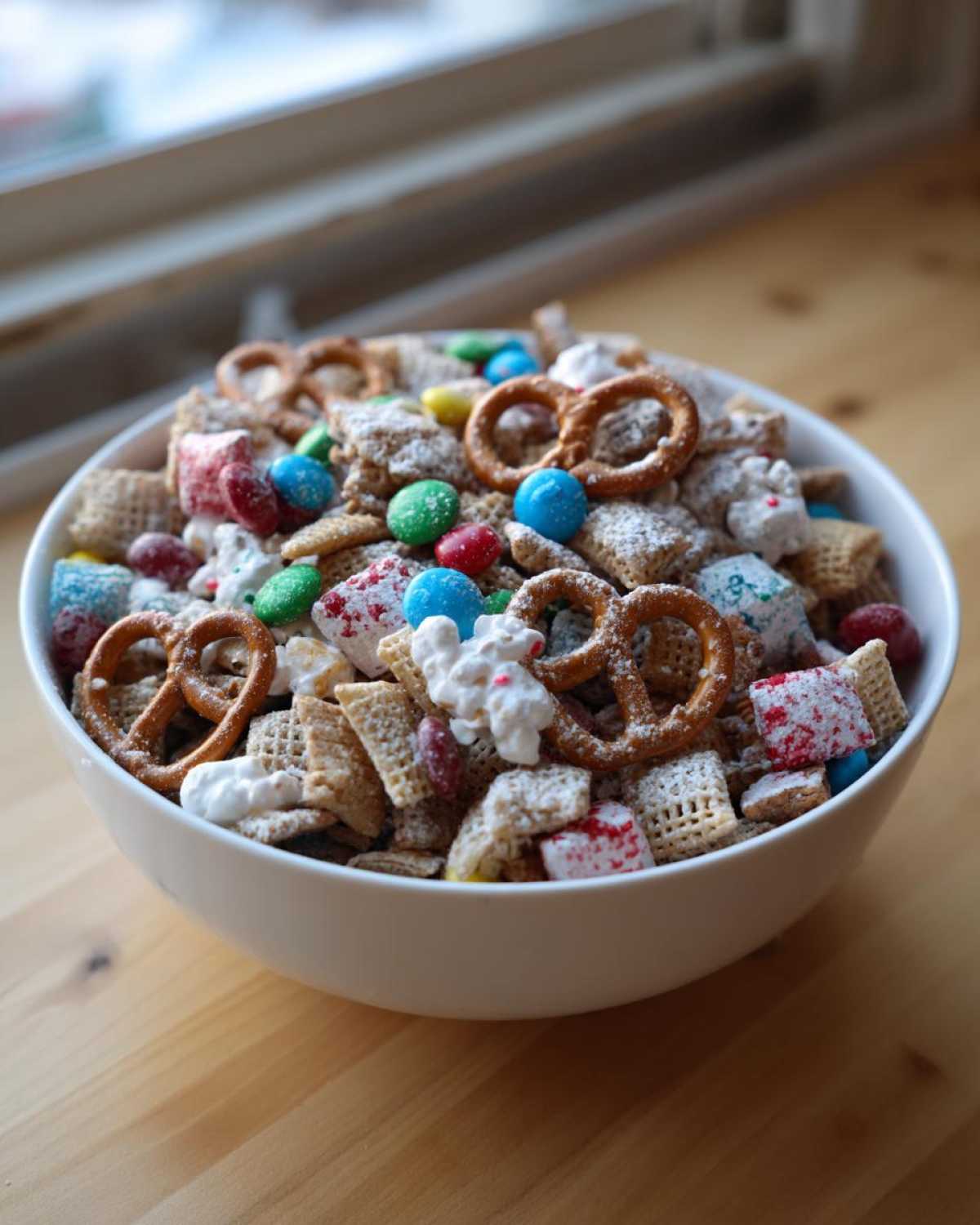 A white bowl filled with a colorful Christmas snack mix featuring pretzels, candies, and cereal pieces, dusted with powdered sugar.