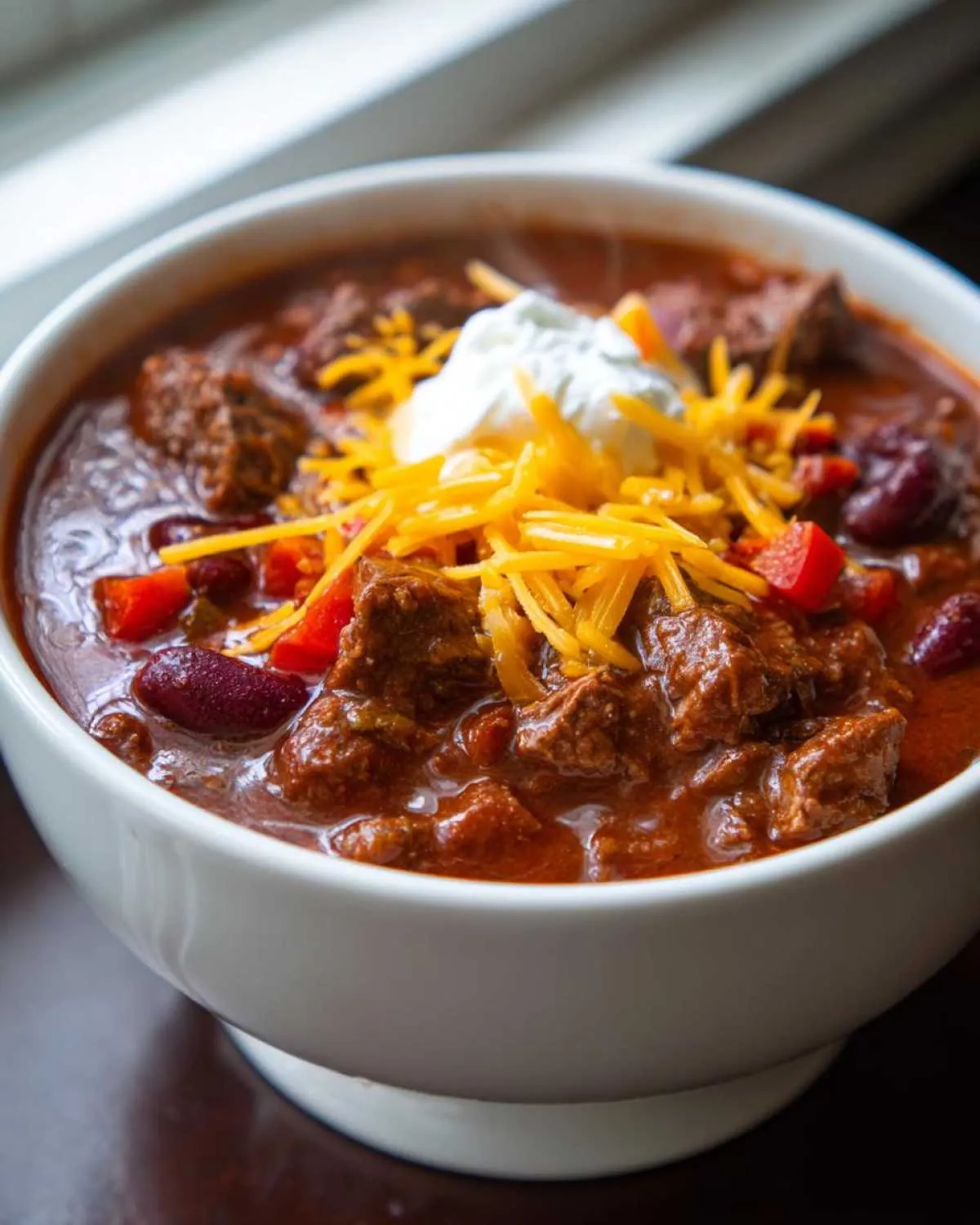 A close-up of a bowl of hearty chuck roast chili, topped with shredded cheese and a dollop of sour cream.