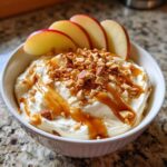 A bowl of creamy cream cheese caramel apple dip topped with chopped nuts, caramel drizzle, and apple slices.