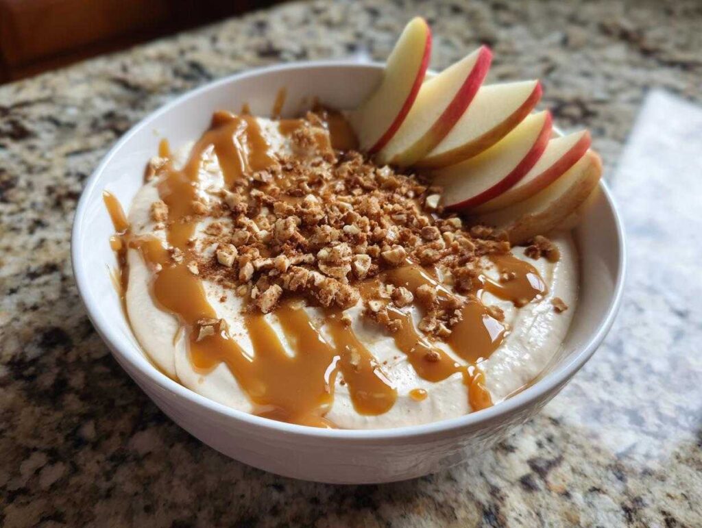 A bowl of creamy cream cheese caramel apple dip topped with caramel drizzle, chopped nuts, and apple slices.