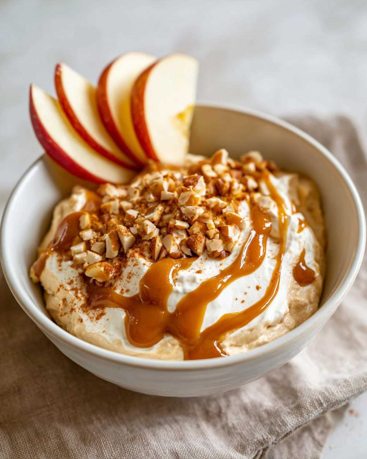 A bowl of creamy cream cheese caramel apple dip topped with whipped cream, chopped nuts, and caramel drizzle, garnished with apple slices.