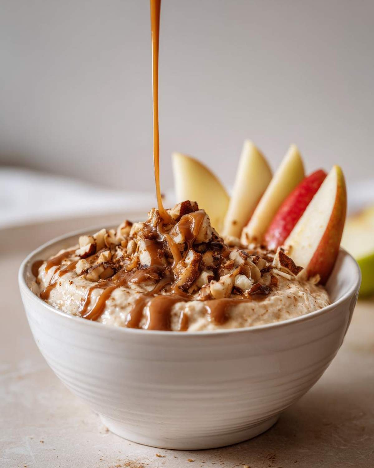 A bowl of Cream Cheese Caramel Apple Dip being drizzled with caramel sauce and topped with chopped nuts, served with apple slices.