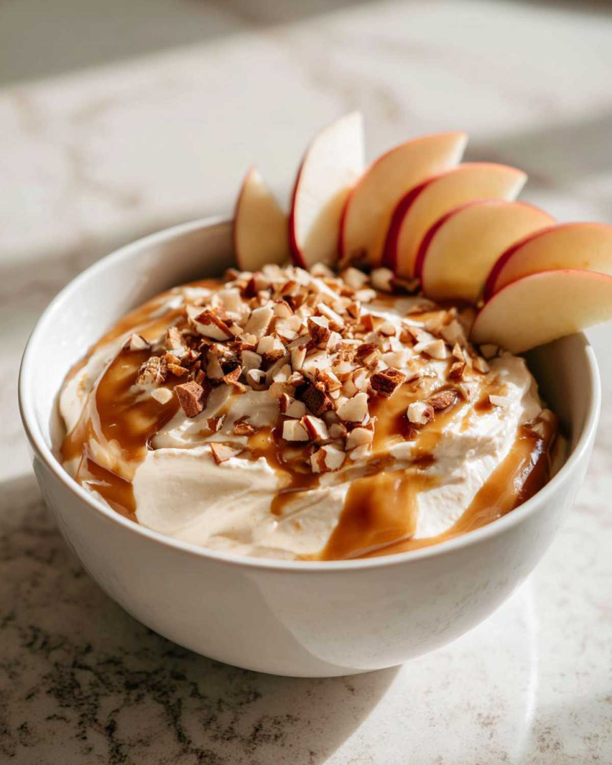 A white bowl filled with creamy cream cheese caramel apple dip, topped with chopped nuts and drizzled with caramel sauce, garnished with apple slices.