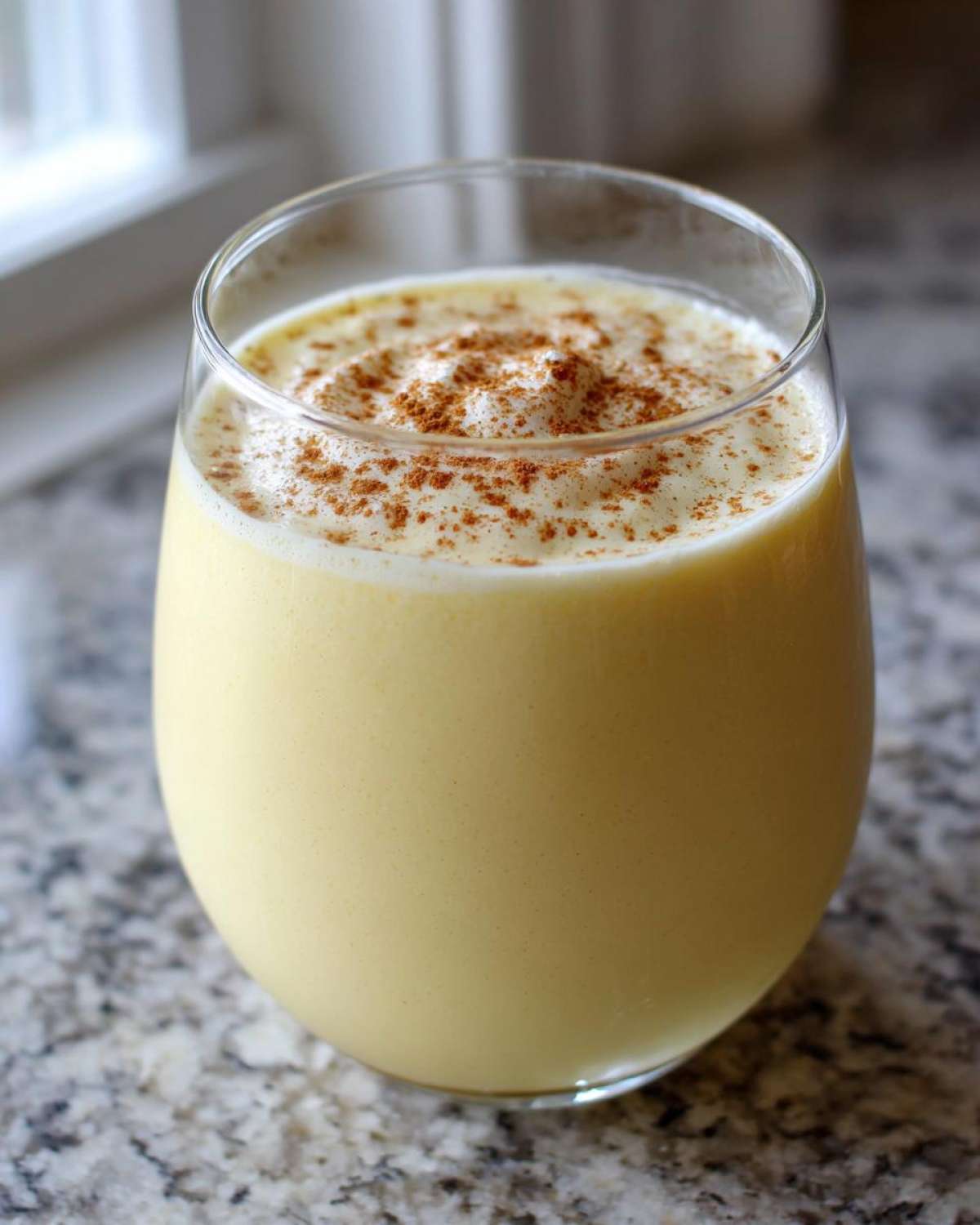 A glass of thick, creamy yellow eggnog topped with foamy cream and a sprinkle of cinnamon.