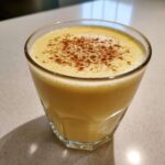 A glass of rich, yellow homemade eggnog topped with a sprinkle of cinnamon.