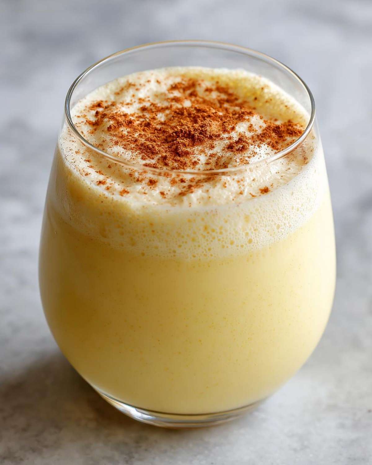 A glass filled with rich, creamy yellow eggnog topped with a dusting of ground cinnamon.
