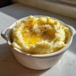A close-up of a bowl of fluffy mashed potatoes topped with melted butter and cracked black pepper.