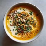 A bowl of creamy roasted pumpkin soup topped with swirls of cream, pumpkin seeds, and fresh herbs.