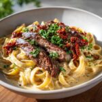 A close-up of creamy steak pasta topped with sliced steak, sun-dried tomatoes, and fresh parsley.