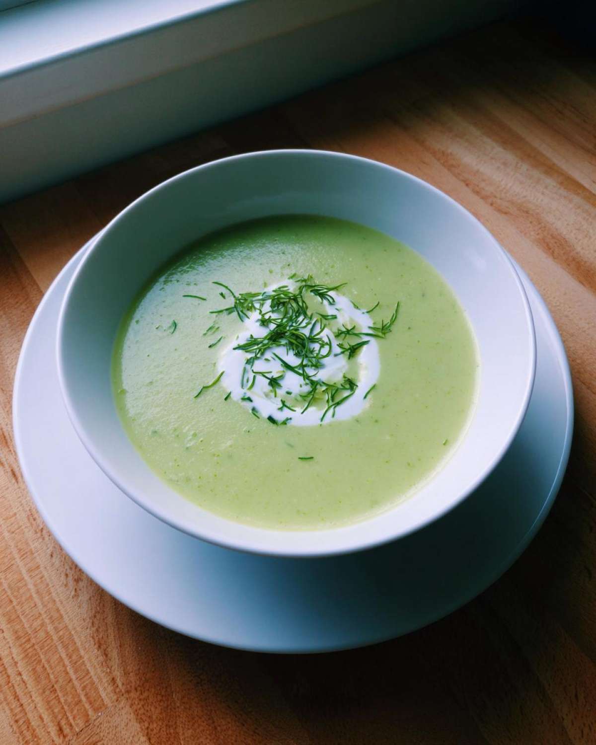 A bowl of smooth, light green zucchini soup topped with a swirl of cream and fresh dill or chives.