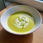 A bowl of vibrant green zucchini soup, garnished with a swirl of cream and fresh green herbs.