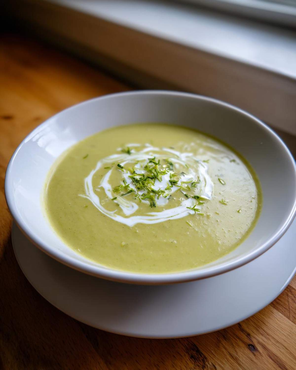 A bowl of smooth, light green zucchini soup, garnished with a swirl of cream and fresh lime zest.