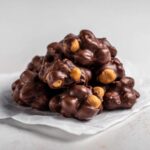 A close-up pile of rich, dark chocolate Crockpot candy clusters studded with peanuts.