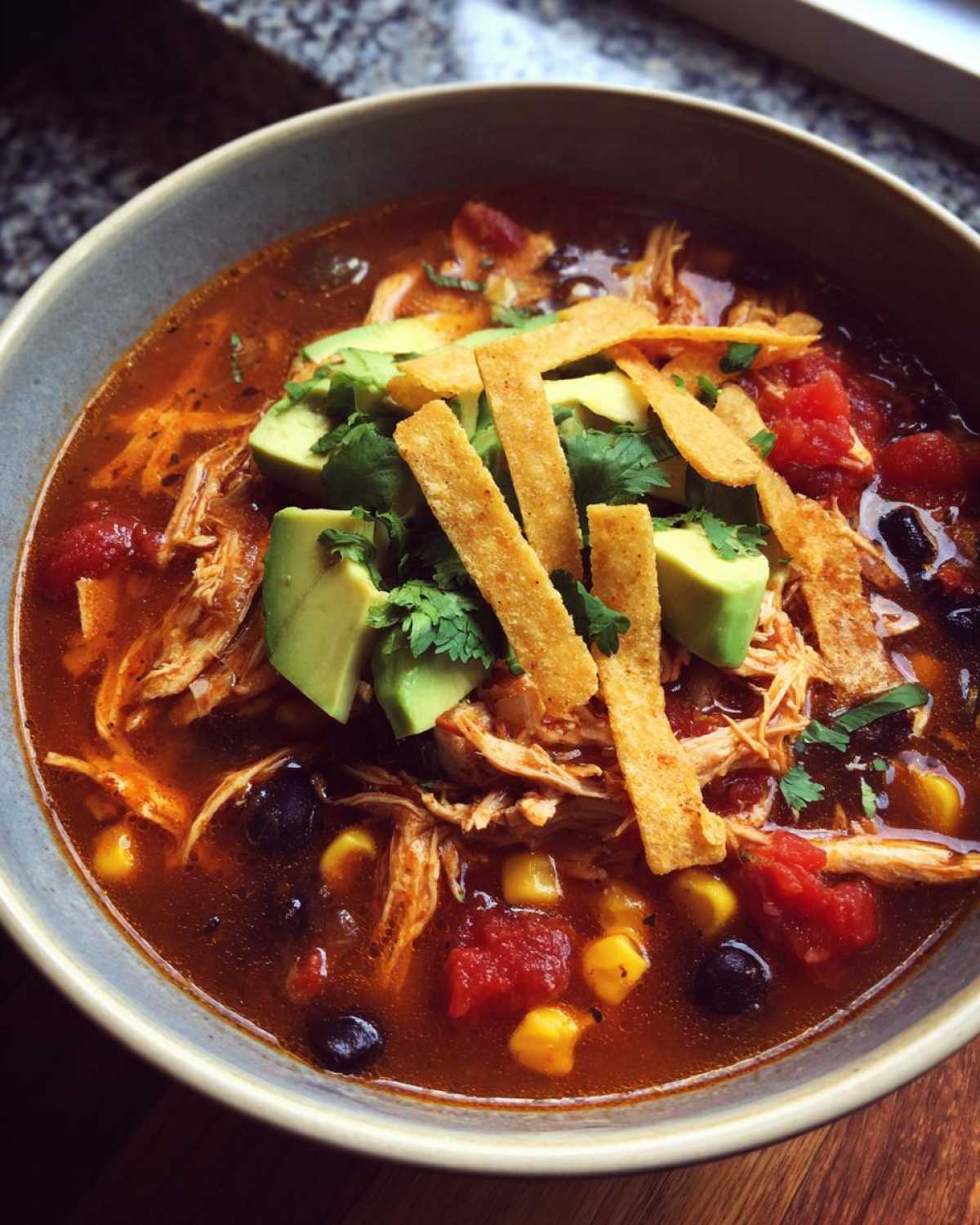 A close-up bowl of crockpot chicken tortilla soup topped with avocado, cilantro, and tortilla strips.