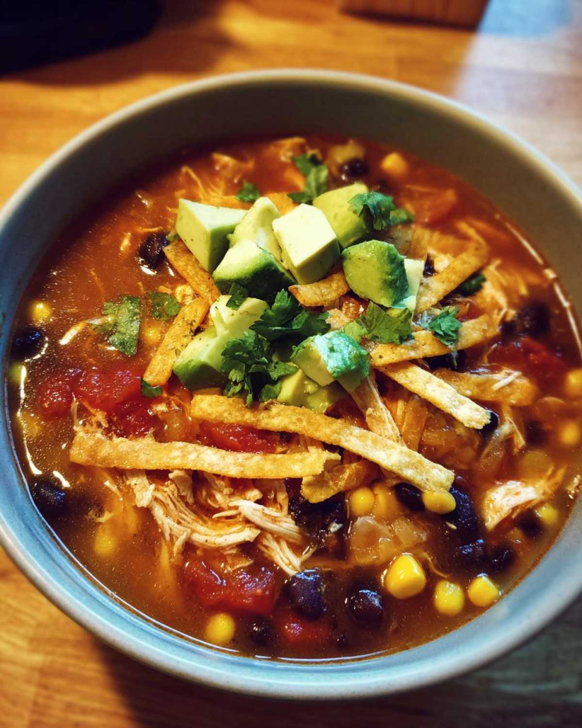 A close-up of a bowl of crockpot chicken tortilla soup, topped with avocado, cilantro, and tortilla strips.