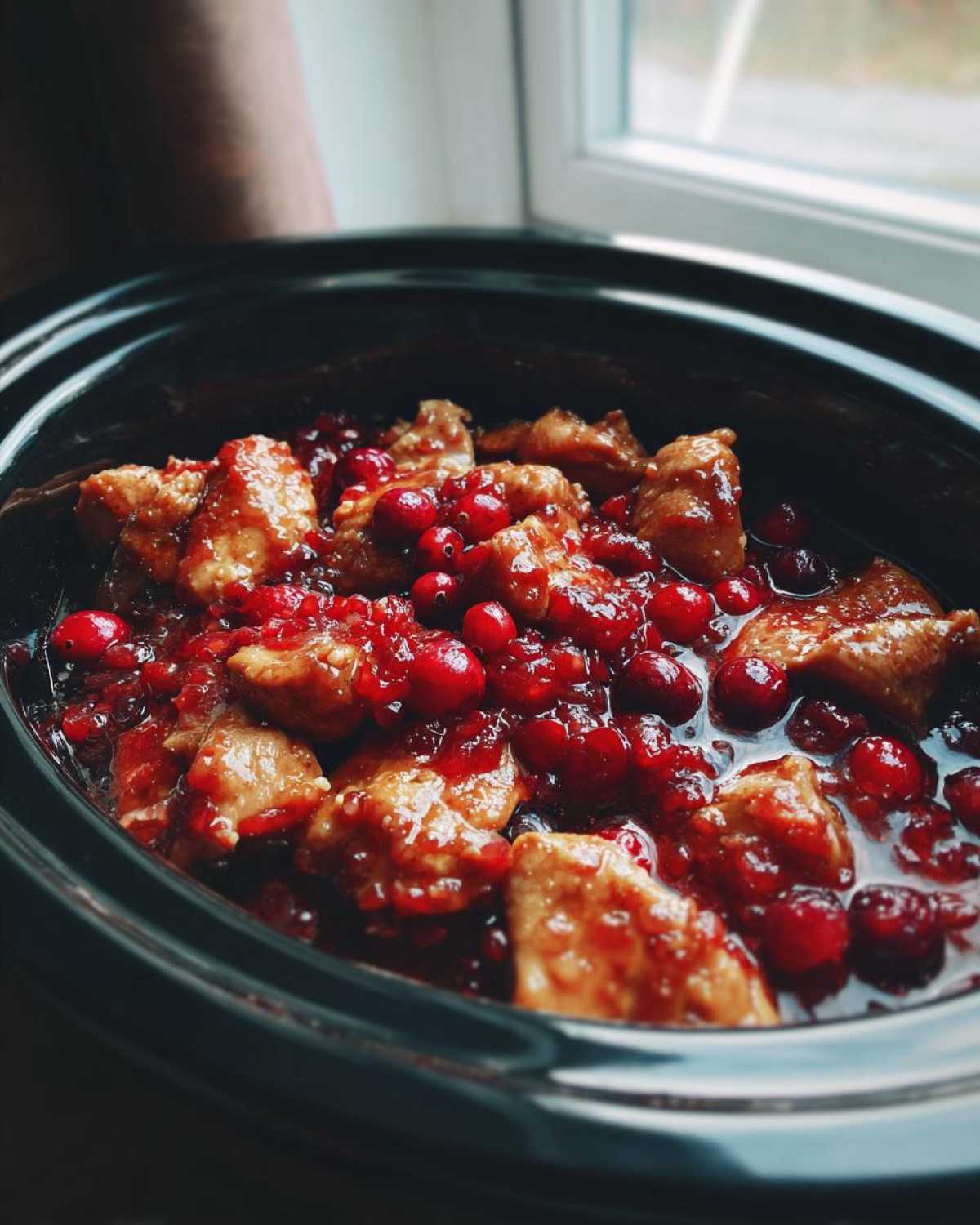 Close-up of tender chicken pieces coated in a glossy cranberry sauce in a black crockpot.
