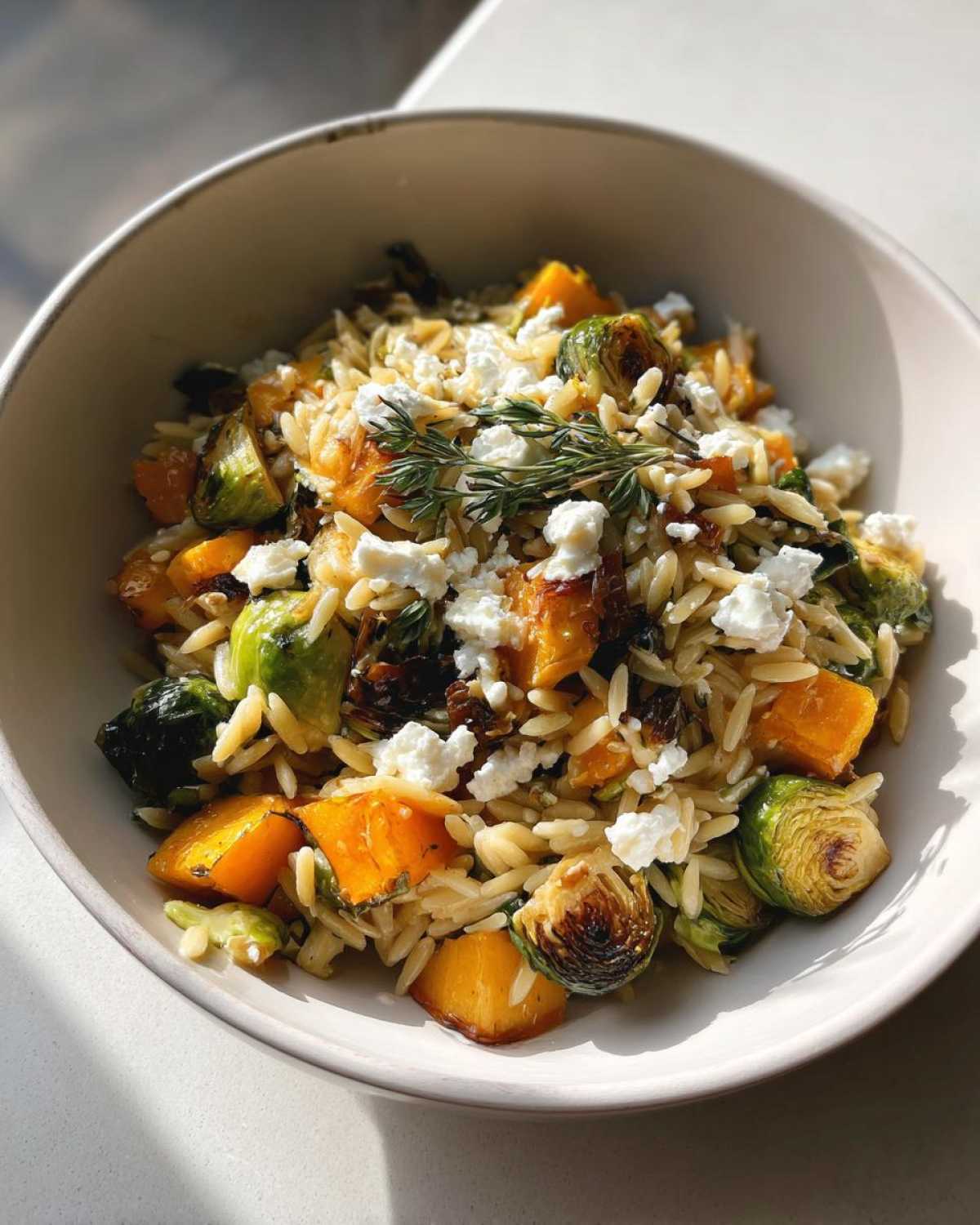 A bowl of fall orzo salad featuring roasted butternut squash, Brussels sprouts, and crumbled feta cheese, garnished with herbs.