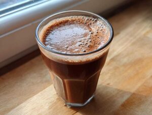 A glass of rich, dark chocolate milk with a foamy top, sitting on a wooden surface near a window.