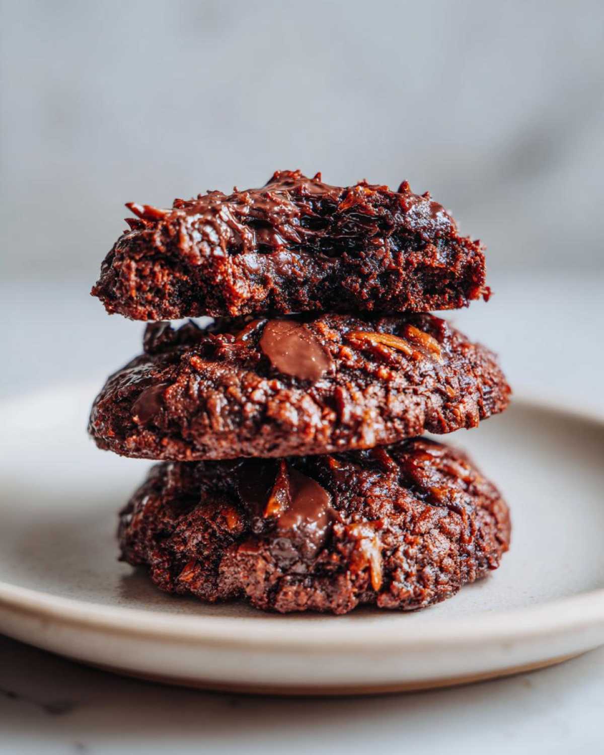 A stack of three dark, fudgy almond joy cookies piled on a light plate, showing melted chocolate chips.