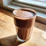 A tall glass filled with frothy, rich chocolate milk sitting on a sunlit wooden surface.