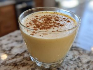 A close-up of a glass filled with creamy, pale yellow eggnog, dusted generously with ground cinnamon.