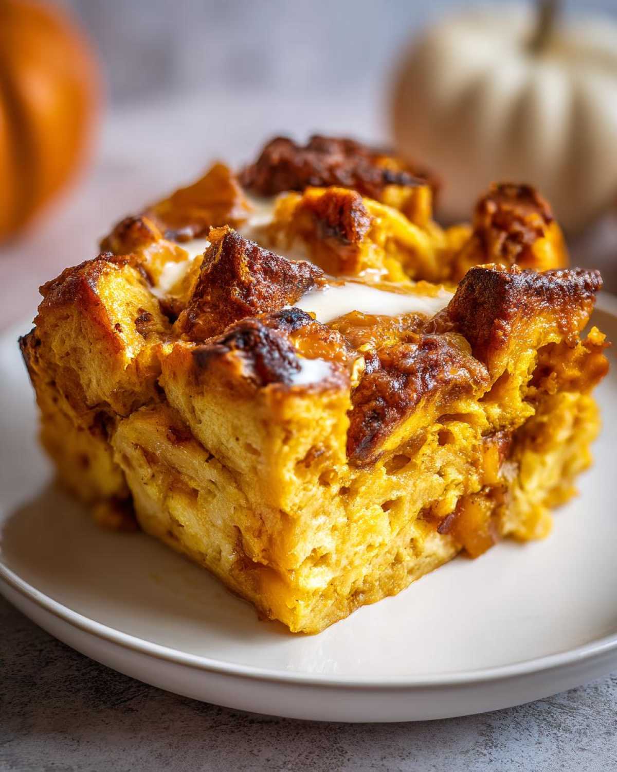 A close-up of a slice of gluten-free pumpkin bread pudding on a white plate, with a creamy sauce drizzled on top.