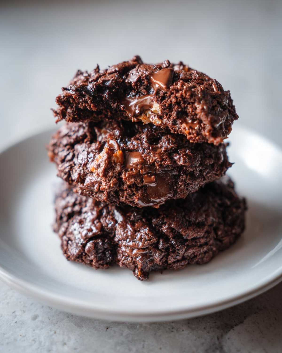 A stack of three rich, dark chocolate Almond Joy cookies, with the top one broken open showing melted chocolate and filling.