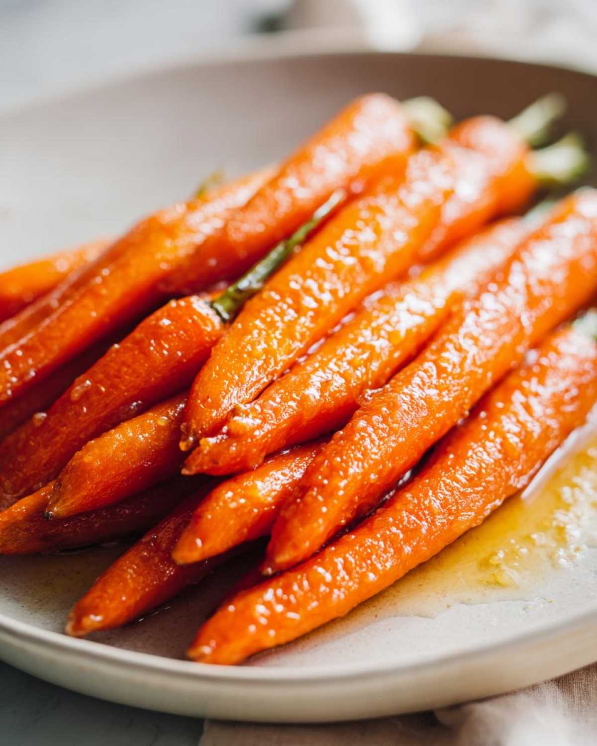 Close-up of bright orange, glossy honey glazed carrots piled on a light-colored plate with visible glaze.