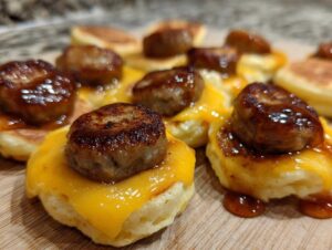 Close-up of several assembled mcgriddle bites featuring mini pancakes, melted cheese, and sausage patties drizzled with syrup.