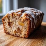 A close-up of a moist apple cake loaf, sliced to show chunks of apple inside, topped with a white vanilla glaze drizzle.