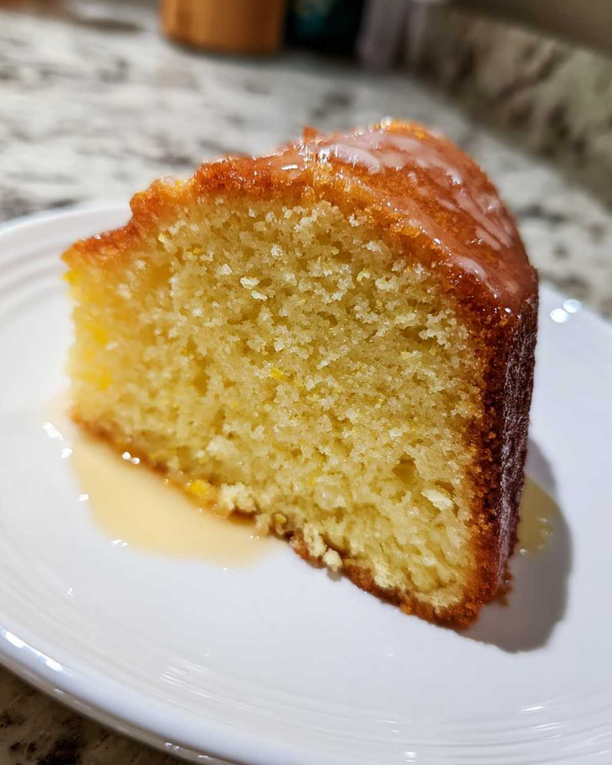 Close-up of a moist slice of olive oil cake with a thin citrus glaze dripping onto a white plate.