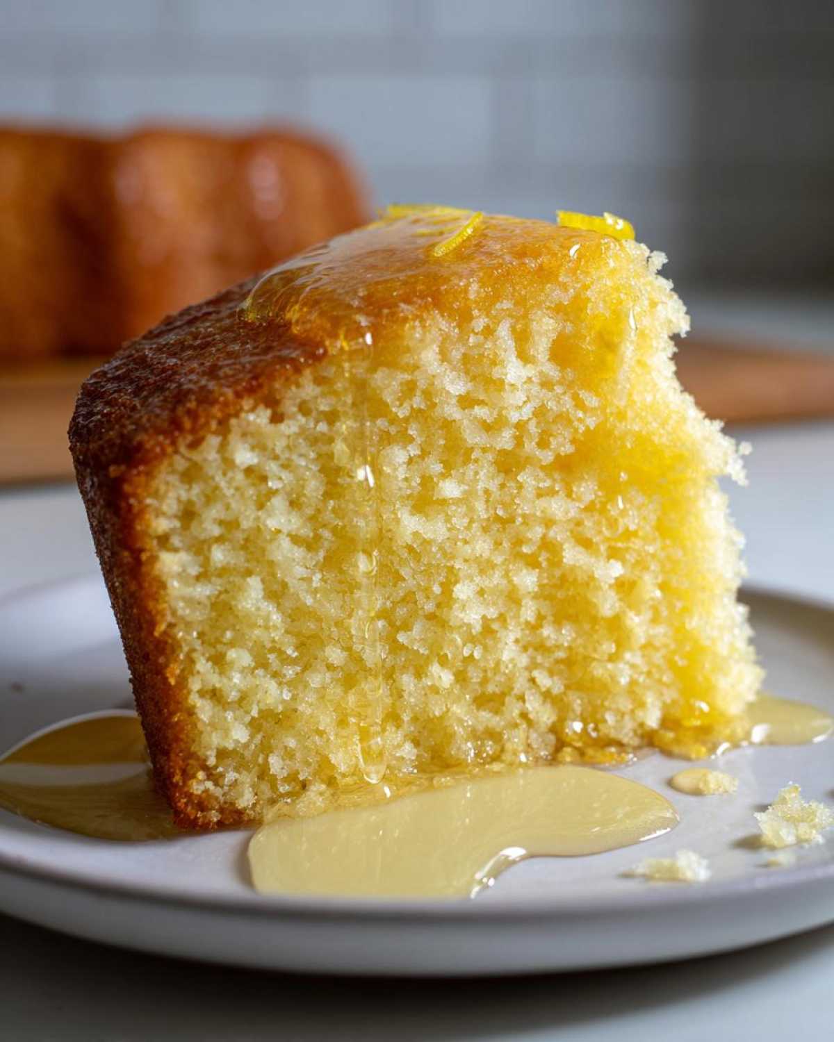 Close-up of a moist slice of yellow olive oil cake drizzled with syrup and topped with lemon zest.