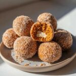 A pile of no bake pumpkin cheesecake balls coated in cinnamon sugar, with one cut open to reveal a creamy white center.