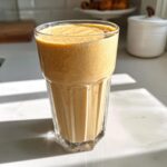 A tall, textured glass filled with a thick, frothy peanut butter banana smoothie sitting on a bright marble countertop.