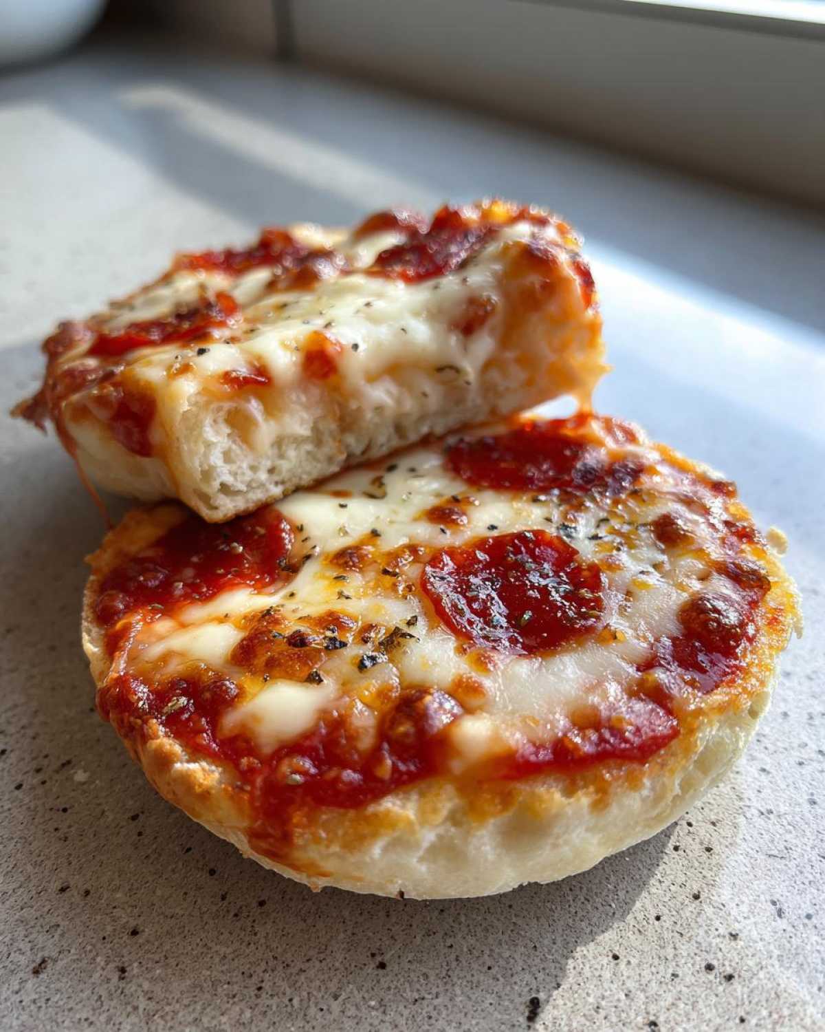 Close-up of a pepperoni english muffin pizza, cut in half, showing melted cheese and herbs.