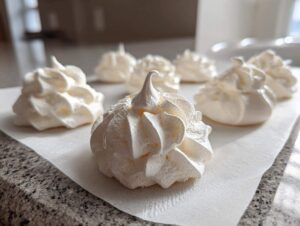 Close-up of freshly piped, white meringue cookies sitting on parchment paper on a kitchen counter.