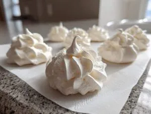 Close-up of freshly piped, white meringue cookies sitting on parchment paper on a kitchen counter.